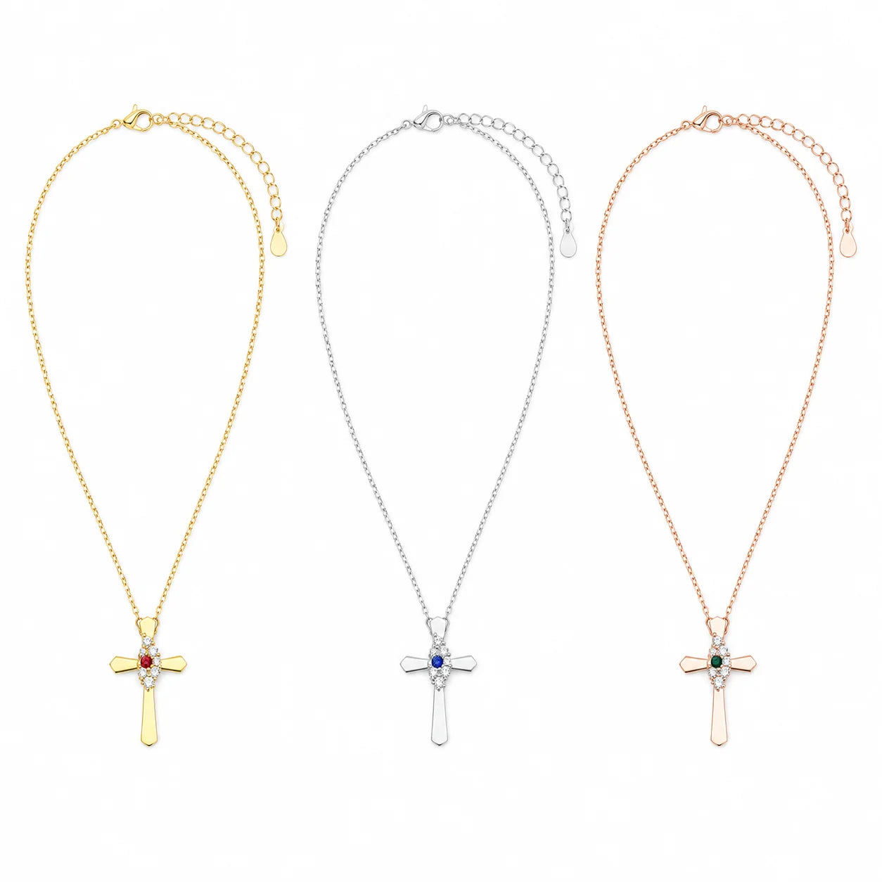 Three cross pendant necklaces shown side by side on a white background, each with a delicate chain and adjustable clasp. The left necklace is gold with a small red gemstone, the center is silver with a blue gemstone, and the right is rose gold with a green gemstone.