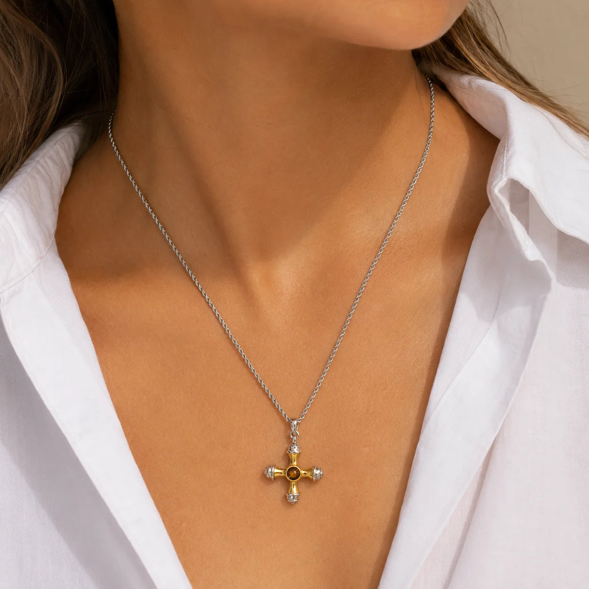 A close-up of a model wearing a silver chain necklace with a cross pendant featuring a gold center and clear gemstones.