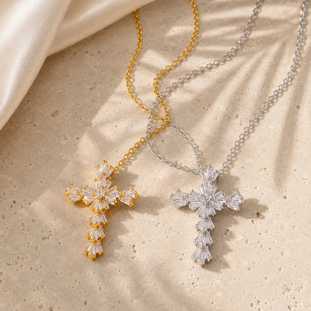 Two cross pendant necklaces with floral starburst designs, one in yellow gold and one in silver, both set with clear crystals, displayed on a textured beige stone surface with a white satin cloth in the corner.