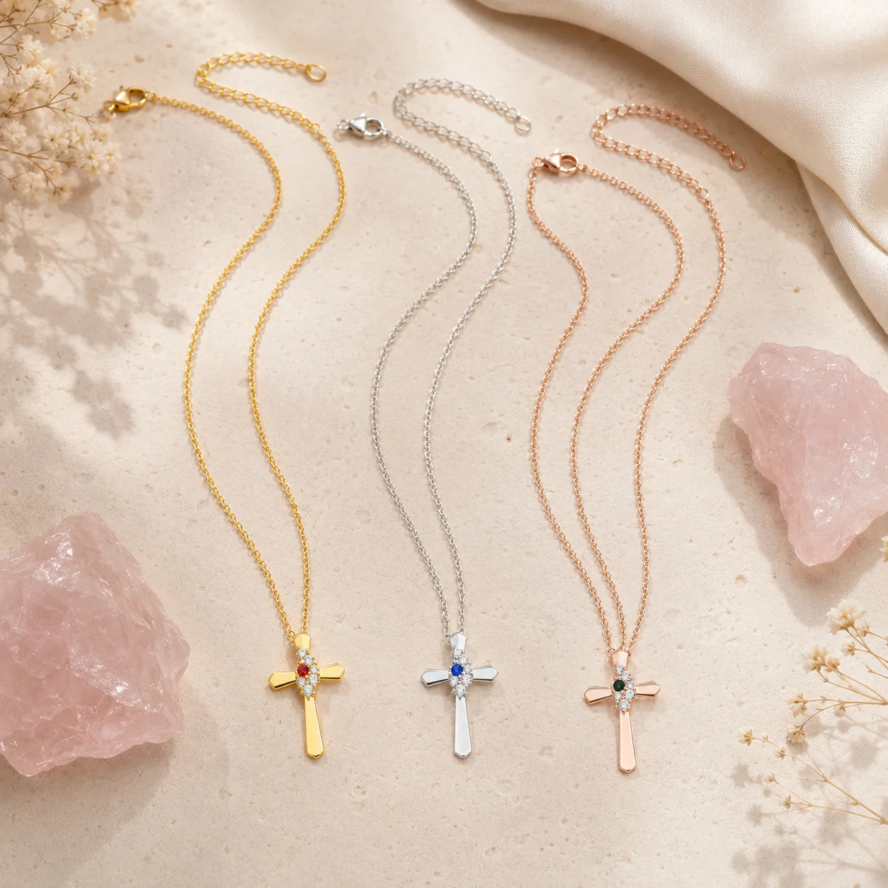 Three delicate cross pendant necklaces in gold, silver, and rose gold are displayed on a light beige textured surface with pink rose quartz crystals and dried flowers. The gold necklace has a small red gemstone, the silver necklace has a blue gemstone, and the rose gold necklace has a green gemstone.