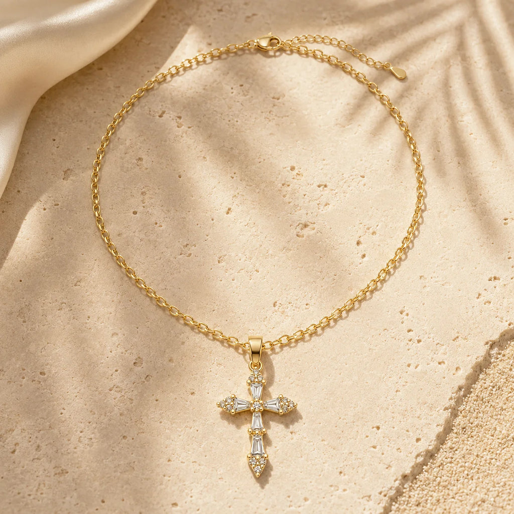 A gold cross pendant necklace with clear crystals, displayed on a textured beige surface with soft shadows.